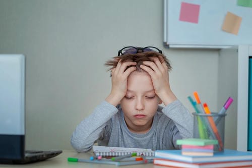A boy sits at a desk surrounded by notebooks, pens and a laptop, clutching his head with both hands, feeling overwhelmed and anxious about his school assignments.