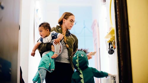 A mother carries one baby in her arms and keeps an eye on her other toddler dressed in a dinosaur onesie as the child explores the hallway, managing care for two children at home.