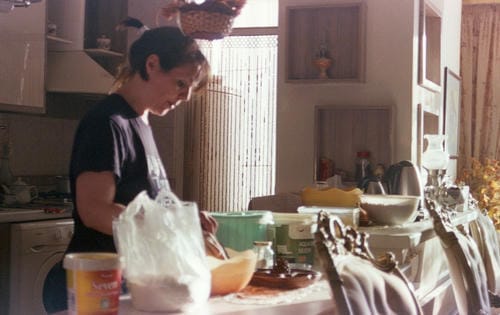 A woman sorts groceries and ingredients on a cluttered kitchen table, busy with daily cooking and domestic preparation work.