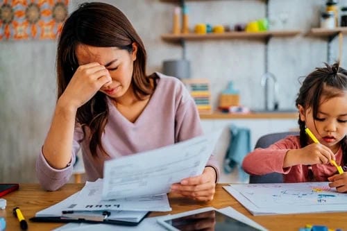 A tired mother presses her forehead with one hand, overwhelmed by piles of bills and documents at the table while her little daughter calmly draws beside her, showing burnout from mixed childcare and household financial stress.