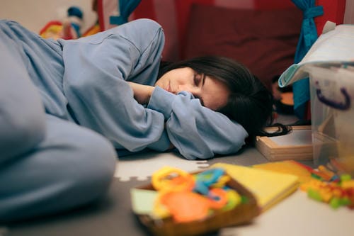 A tired mother lies fast asleep on the floor surrounded by scattered children’s toys, falling into slumber after long hours of looking after her kid.