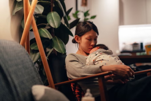 A worn-out mother falls asleep in an armchair with her sleeping infant held safely in her arms, drained from round-the-clock childcare work.